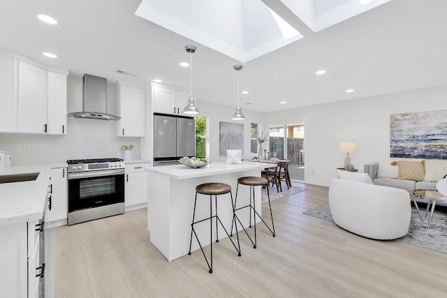 kitchen with appliances with stainless steel finishes, white cabinetry, hanging light fixtures, a center island, and wall chimney exhaust hood