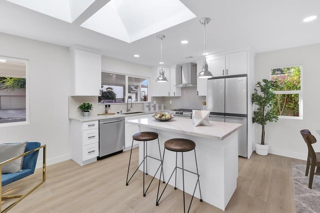 kitchen with pendant lighting, appliances with stainless steel finishes, white cabinetry, a kitchen island, and wall chimney exhaust hood