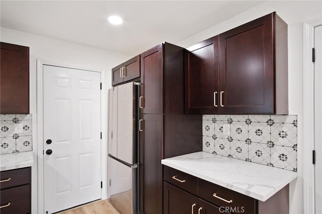 kitchen with dark brown cabinets, fridge, light stone counters, light hardwood / wood-style floors, and decorative backsplash