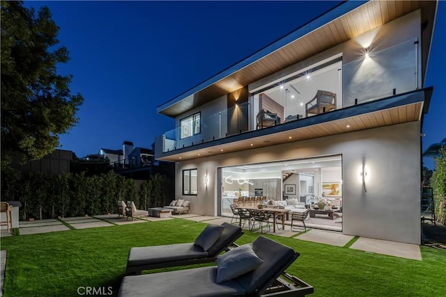 back house at night with an outdoor living space, a patio, a balcony, and a lawn