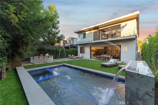 back house at dusk with a balcony, a lawn, exterior bar, pool water feature, and a patio area