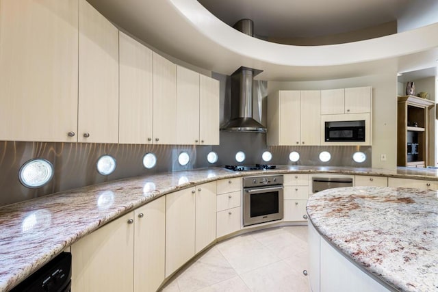 kitchen featuring light stone counters, black appliances, wall chimney range hood, cream cabinets, and backsplash