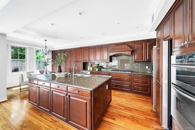 kitchen featuring pendant lighting, sink, premium range hood, stainless steel appliances, and an island with sink