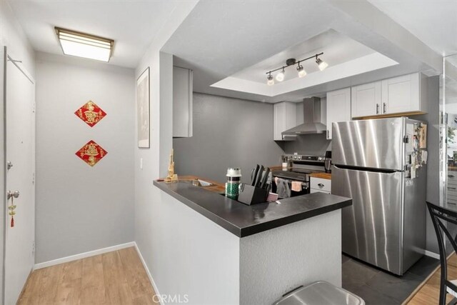 kitchen with wall chimney exhaust hood, white cabinetry, light wood-type flooring, a raised ceiling, and stainless steel appliances