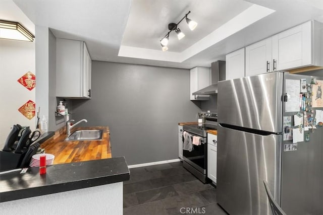 kitchen featuring sink, wall chimney range hood, a tray ceiling, stainless steel appliances, and white cabinets