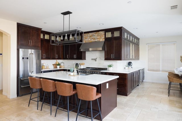 kitchen with stainless steel appliances, a kitchen island with sink, dark brown cabinets, and wall chimney range hood