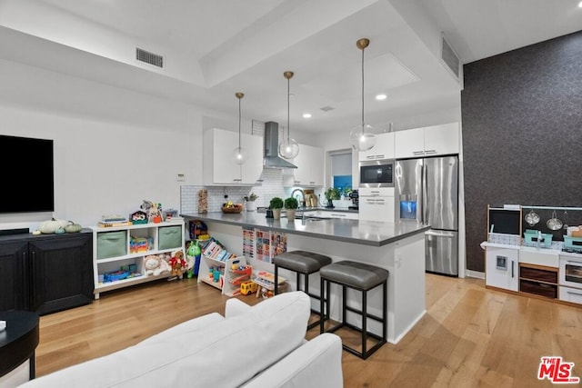 kitchen featuring appliances with stainless steel finishes, decorative light fixtures, white cabinets, a kitchen breakfast bar, and wall chimney range hood