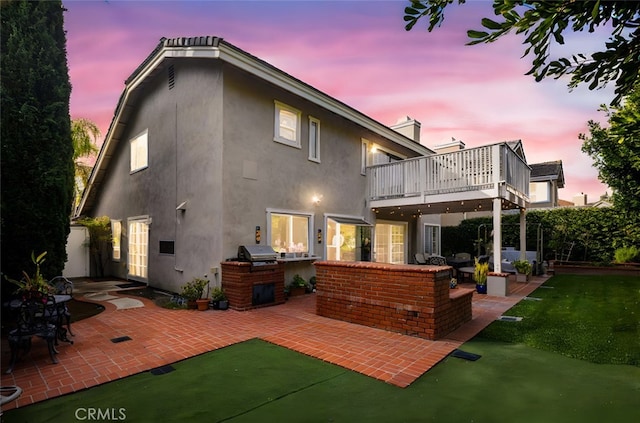 back house at dusk with a balcony, a patio area, and a lawn