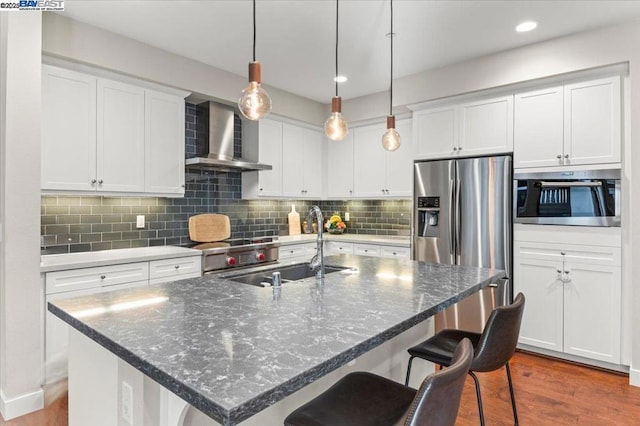 kitchen with wall chimney range hood, sink, a breakfast bar area, white cabinetry, and a center island with sink