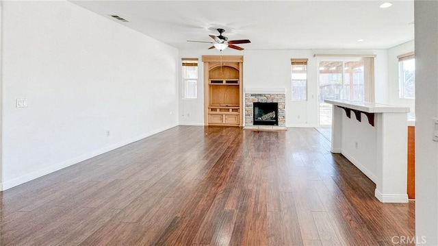 unfurnished living room with a stone fireplace, dark wood-type flooring, and ceiling fan