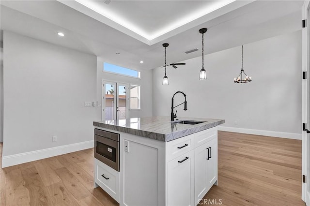 kitchen featuring stainless steel microwave, an island with sink, sink, white cabinets, and hanging light fixtures