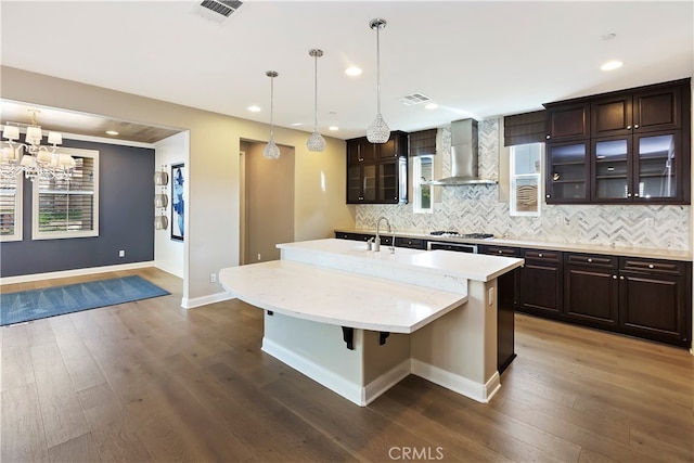 kitchen featuring dark brown cabinetry, a kitchen breakfast bar, a kitchen island with sink, decorative backsplash, and wall chimney range hood