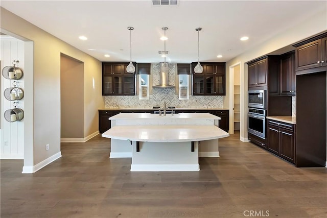 kitchen featuring wall chimney exhaust hood, dark brown cabinets, a center island with sink, stainless steel appliances, and backsplash