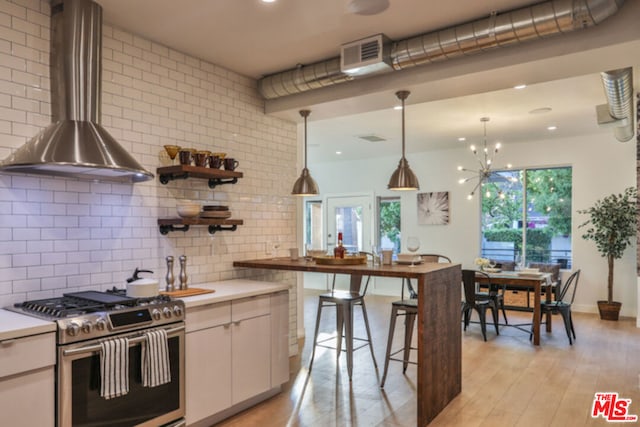 kitchen featuring tasteful backsplash, white cabinets, hanging light fixtures, wall chimney range hood, and stainless steel gas range