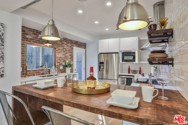 kitchen with sink, white cabinetry, hanging light fixtures, brick wall, and stainless steel fridge with ice dispenser