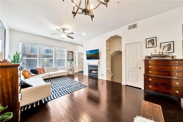 living area with arched walkways, dark wood-style flooring, visible vents, a glass covered fireplace, and ceiling fan with notable chandelier