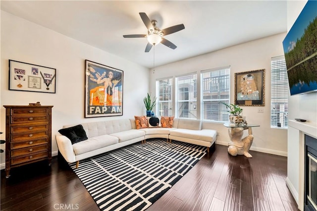 living room featuring ceiling fan, dark wood-type flooring, a glass covered fireplace, and baseboards