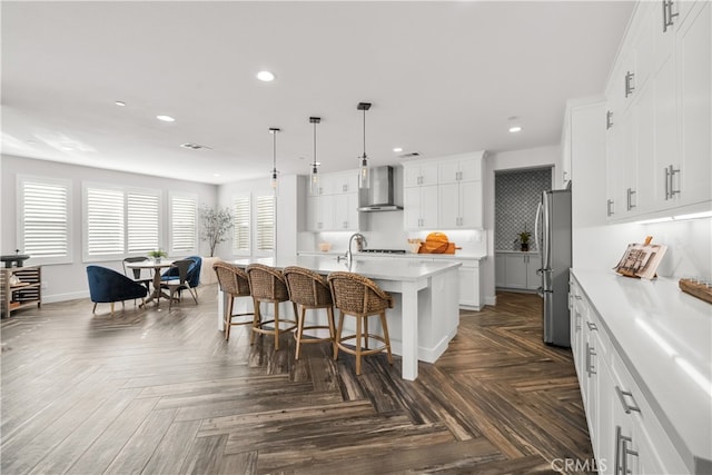 kitchen with pendant lighting, white cabinetry, an island with sink, stainless steel fridge, and wall chimney exhaust hood