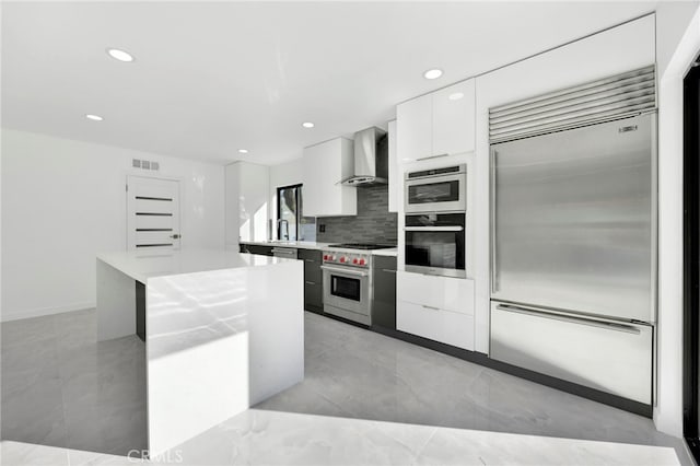 kitchen with white cabinets, premium appliances, decorative backsplash, a center island, and wall chimney range hood