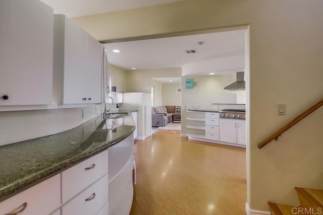 kitchen featuring wall chimney range hood, sink, light hardwood / wood-style flooring, white cabinetry, and dark stone counters