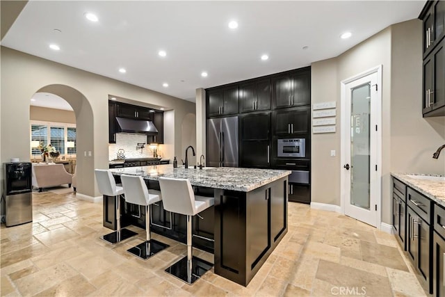 kitchen with light stone counters, a kitchen island with sink, under cabinet range hood, stainless steel fridge, and stone tile flooring