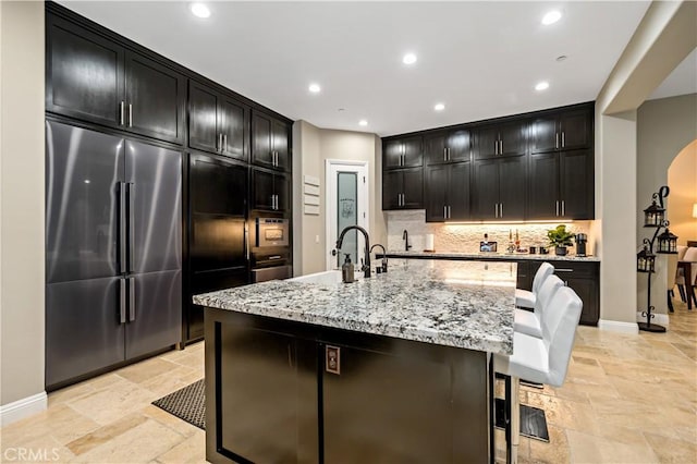 kitchen with a center island with sink, decorative backsplash, light stone countertops, stainless steel built in fridge, and a sink