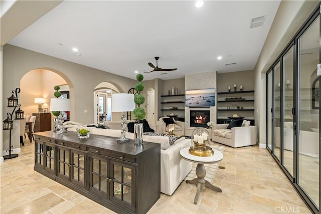 living room featuring arched walkways, stone tile floors, a ceiling fan, visible vents, and a tiled fireplace