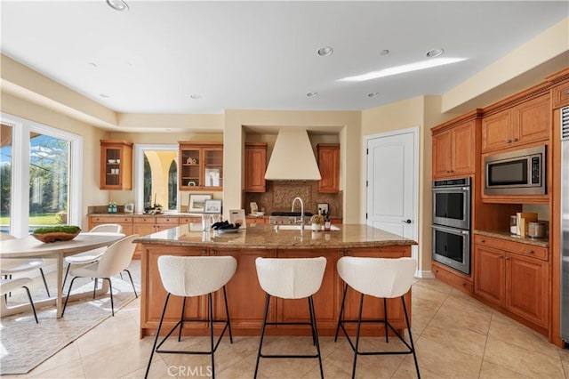 kitchen featuring sink, premium range hood, stainless steel appliances, tasteful backsplash, and an island with sink