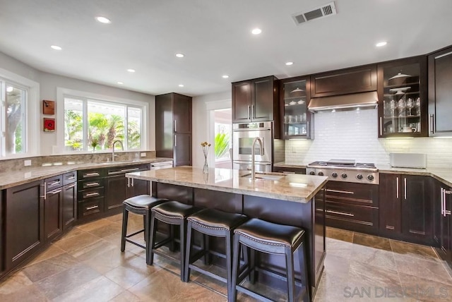 kitchen featuring light stone counters, tasteful backsplash, a kitchen breakfast bar, an island with sink, and stainless steel appliances