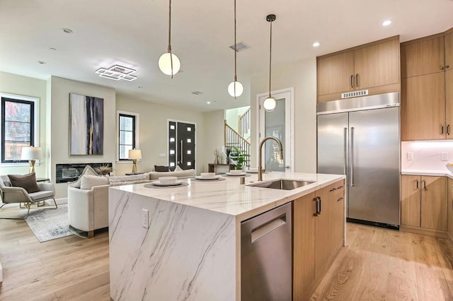 kitchen with sink, light wood-type flooring, an island with sink, stainless steel appliances, and light stone countertops