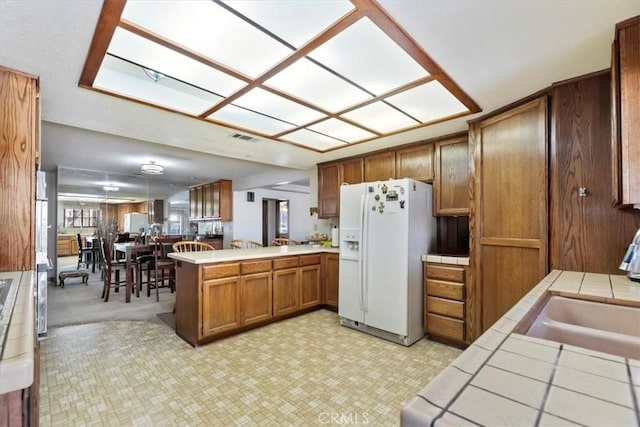 kitchen featuring sink, tile counters, white fridge with ice dispenser, and kitchen peninsula