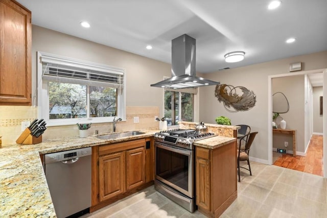 kitchen featuring appliances with stainless steel finishes, island range hood, sink, light stone counters, and kitchen peninsula