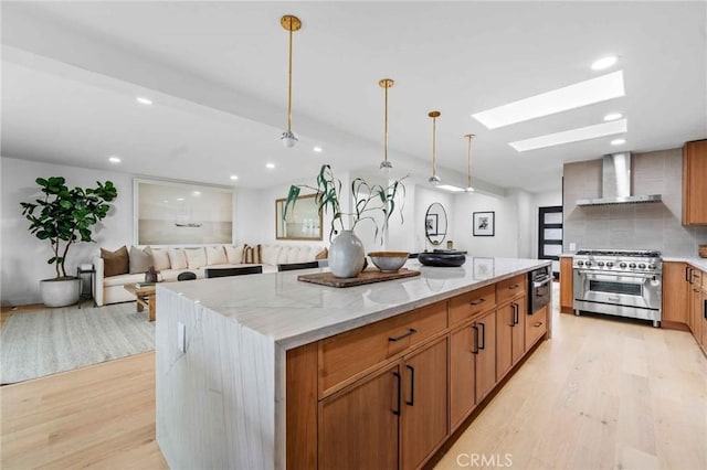 kitchen with a spacious island, wall chimney exhaust hood, hanging light fixtures, light wood-type flooring, and stainless steel stove