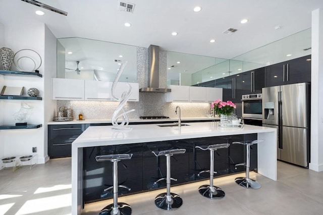 kitchen with appliances with stainless steel finishes, sink, wall chimney range hood, and a kitchen island with sink