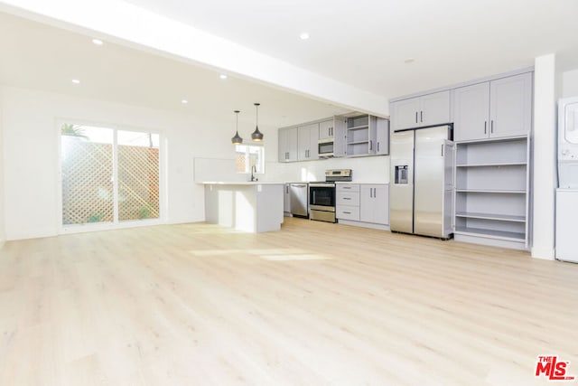 kitchen with appliances with stainless steel finishes, gray cabinetry, hanging light fixtures, stacked washer and clothes dryer, and light wood-type flooring