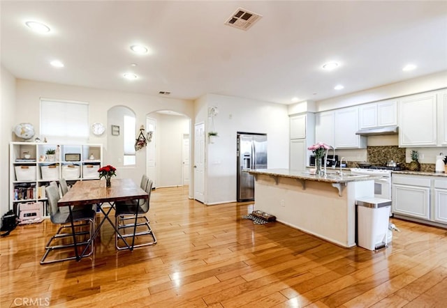 kitchen featuring a breakfast bar, white cabinetry, an island with sink, stainless steel fridge, and light stone counters