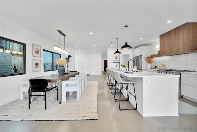 kitchen with appliances with stainless steel finishes, white cabinets, backsplash, hanging light fixtures, and a kitchen island with sink