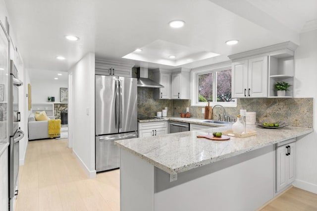 kitchen featuring appliances with stainless steel finishes, white cabinets, kitchen peninsula, light stone countertops, and wall chimney range hood