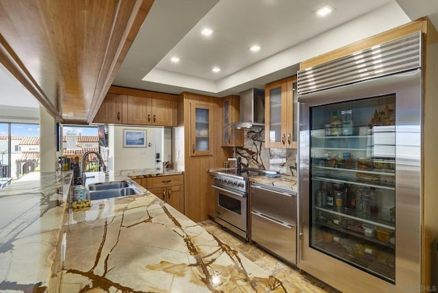 kitchen with sink, high end appliances, a tray ceiling, light stone countertops, and wall chimney exhaust hood