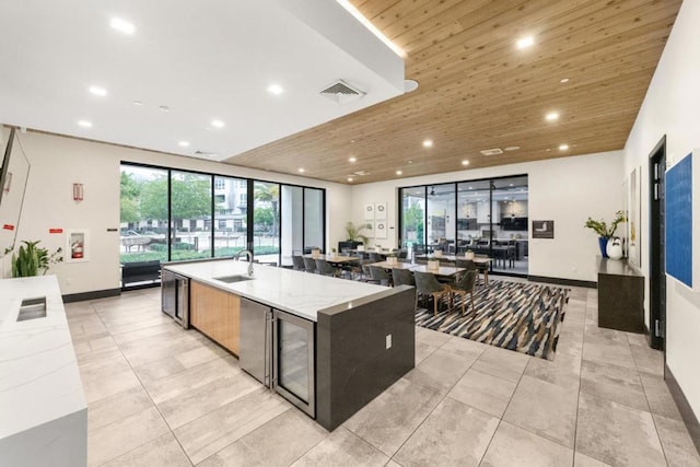 kitchen with sink, wood ceiling, wine cooler, light stone countertops, and a center island with sink