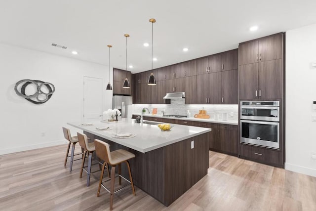 kitchen with dark brown cabinetry, hanging light fixtures, stainless steel appliances, a kitchen island with sink, and decorative backsplash