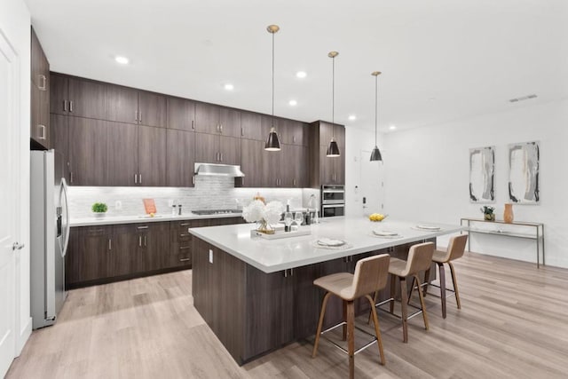 kitchen featuring an island with sink, hanging light fixtures, stainless steel appliances, dark brown cabinets, and light wood-type flooring