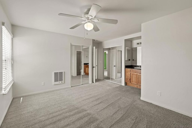 unfurnished bedroom with heating unit, connected bathroom, two closets, carpet, and ceiling fan