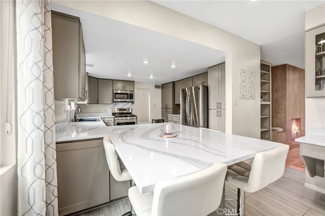 kitchen featuring a kitchen breakfast bar, a peninsula, gray cabinetry, stainless steel appliances, and light stone counters