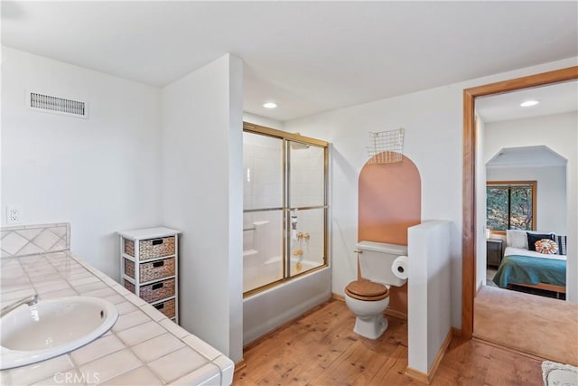 bathroom with bath / shower combo with glass door, visible vents, toilet, a sink, and wood finished floors