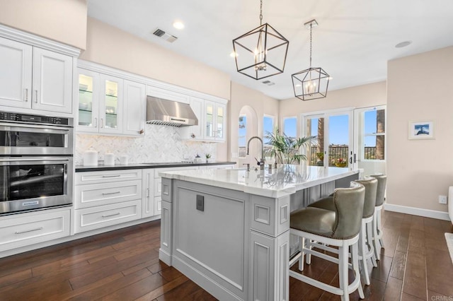 kitchen featuring pendant lighting, double oven, white cabinetry, an island with sink, and wall chimney range hood