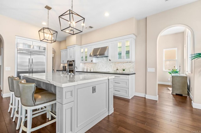 kitchen with dark wood-style floors, arched walkways, decorative backsplash, a kitchen island with sink, and wall chimney exhaust hood