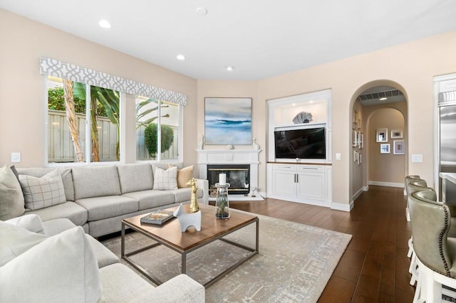 living area with baseboards, arched walkways, a glass covered fireplace, dark wood-style flooring, and recessed lighting