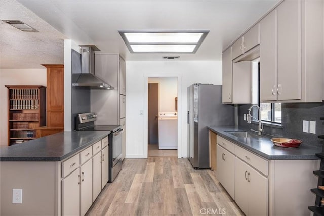 kitchen featuring wall chimney range hood, sink, appliances with stainless steel finishes, light hardwood / wood-style floors, and washer / dryer