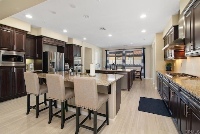 kitchen featuring a breakfast bar, appliances with stainless steel finishes, light stone counters, an island with sink, and wall chimney exhaust hood